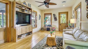 A spacious living room with a large flat-screen TV, wooden shelving, a leather sofa, and a ceiling fan. - Eclectic Mandeville Home: Walk to Lakefront Park (Mandeville)