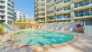 Una piscina con sillones de descanso, rodeada por un edificio de gran altura con balcones.