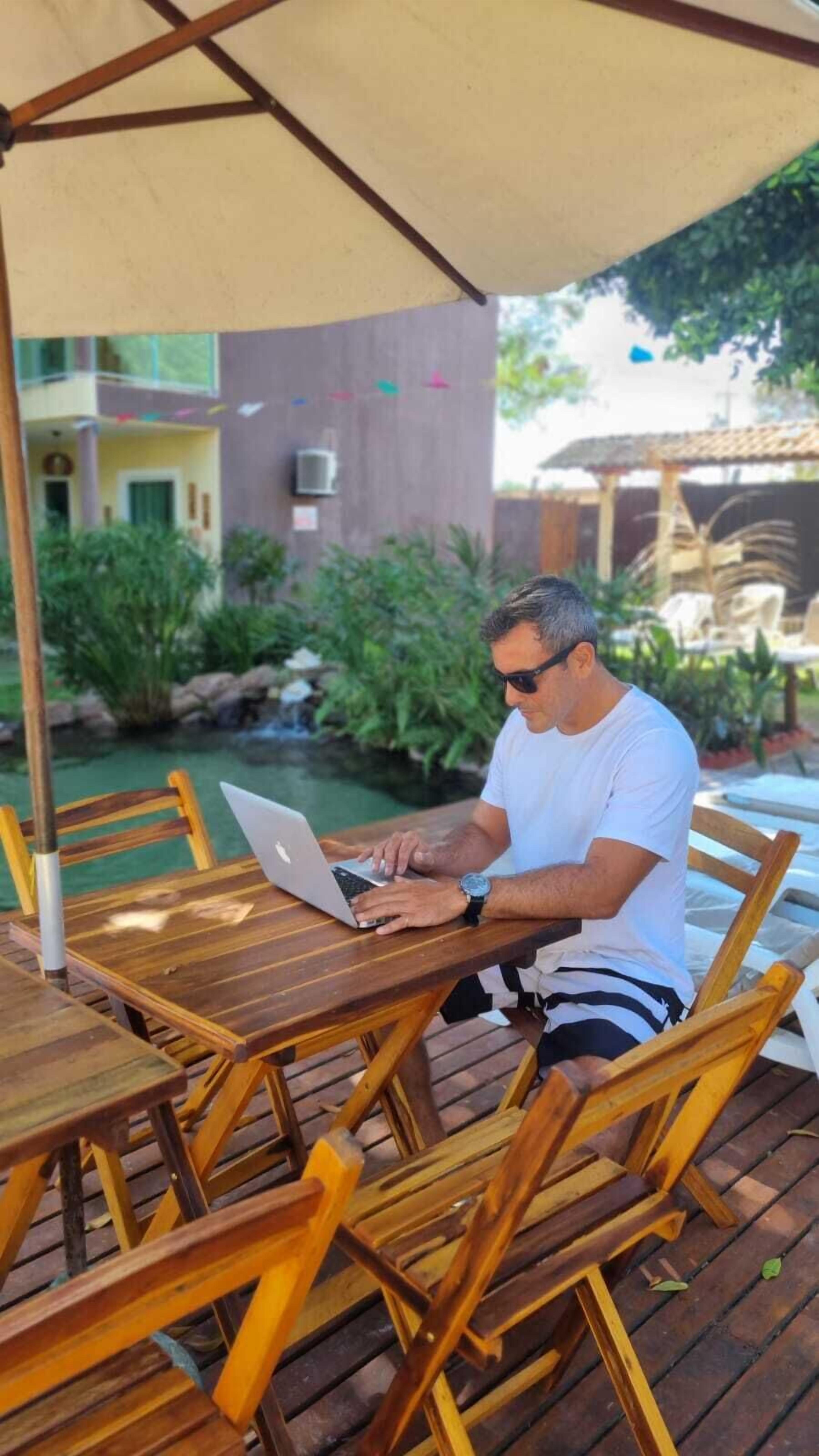 Un hombre sentado en una mesa de madera al aire libre con una computadora portátil, bajo un toldo.