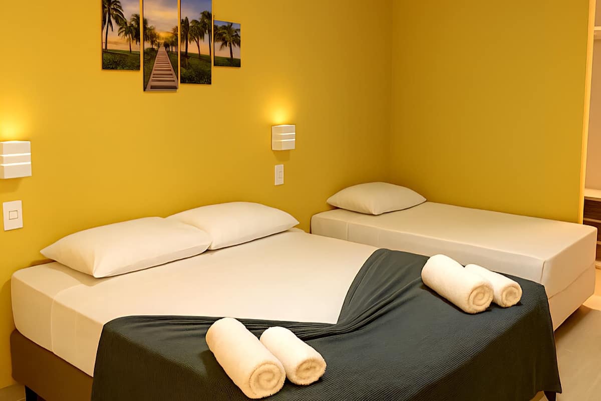 Ein Hotelzimmer mit einem großen Bett, einem kleineren Bett und einer Bank. An den Wänden sind Lampen angebracht und eine Klimaanlage vorhanden.