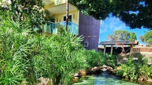 Un estanque cristalino con peces koi, rodeado de exuberante vegetación y un edificio con balcón.