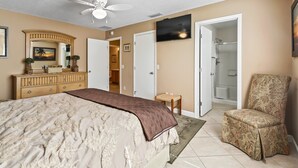 Un dormitorio con cama, ventilador de techo, cómoda, silla, televisor y baño visible a través de una puerta abierta.