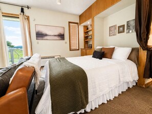 A bedroom with a bed, a bench, a window with curtains, a shelf with decorative items, and a painting on the wall.