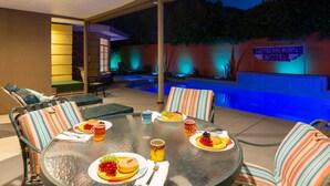 Un espacio de comedor al aire libre con una mesa redonda de vidrio, sillas a rayas y una piscina con iluminación azul.
