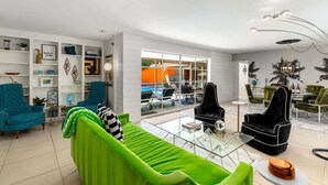 Un salón moderno con un sofá verde, sillones turquesa y un sillón de terciopelo negro. Hay estantes con objetos decorativos y un ventanal con vistas.