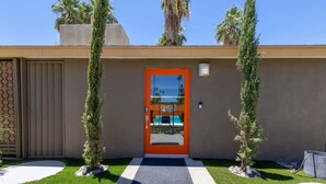 Un edificio moderno con una puerta naranja distintiva, un pequeño jardín con un sendero y una palmera.