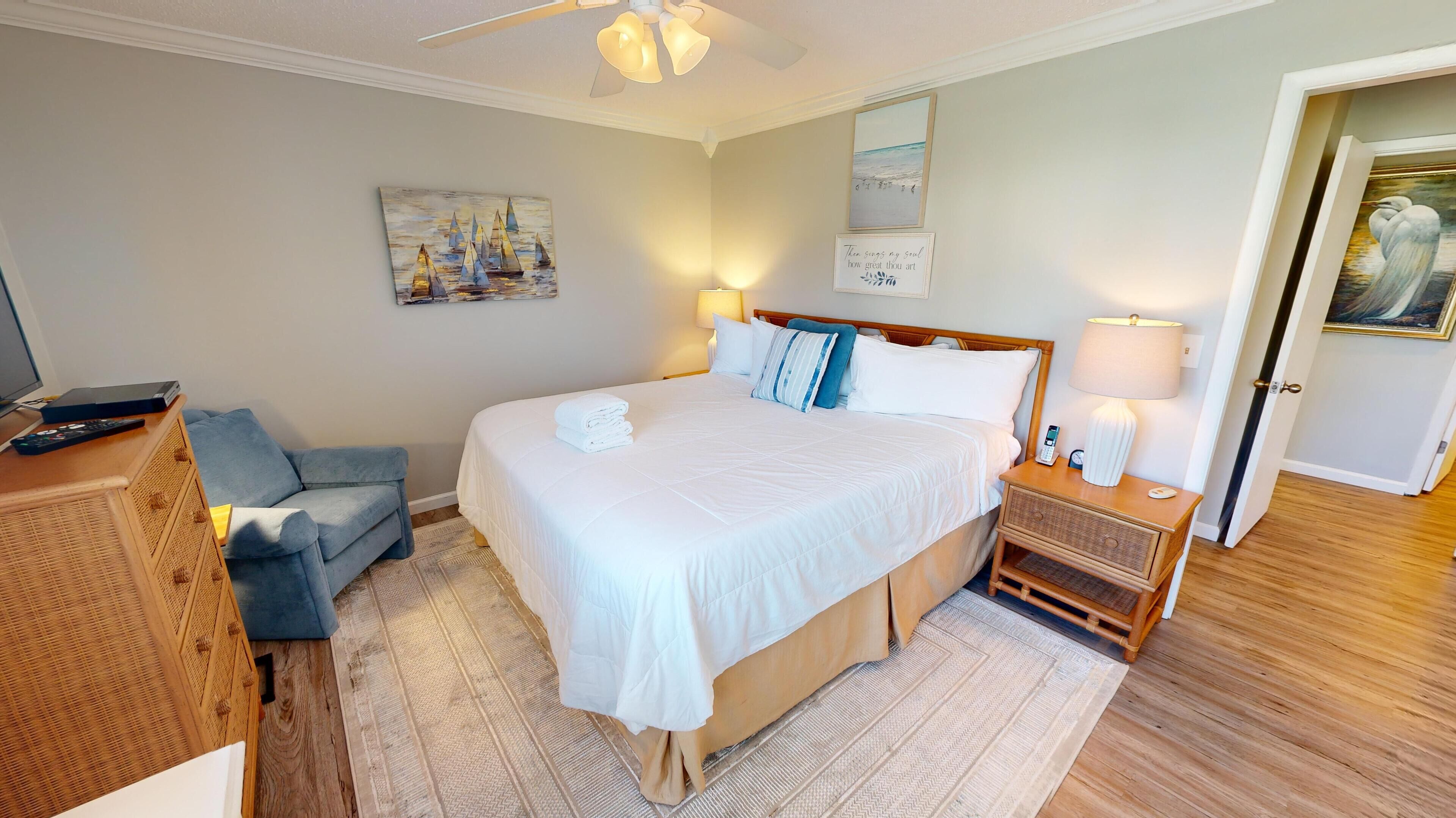 Un dormitorio ordenado con una cama grande, una mesita de noche con lámpara, una silla y un cuadro en la pared.
