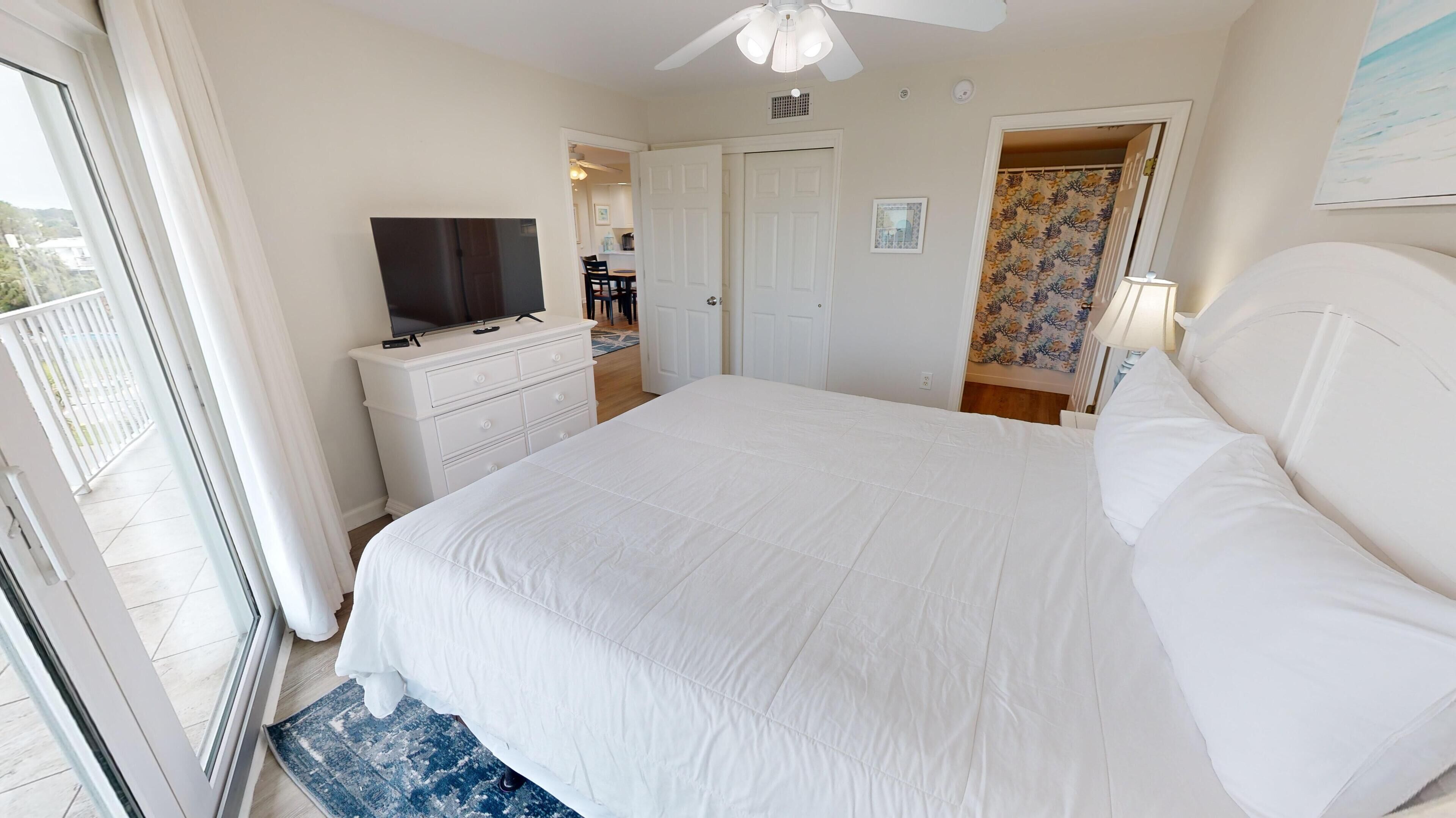 A bedroom with a large bed, a TV on a dresser, a ceiling fan, and a view of the outside balcony.