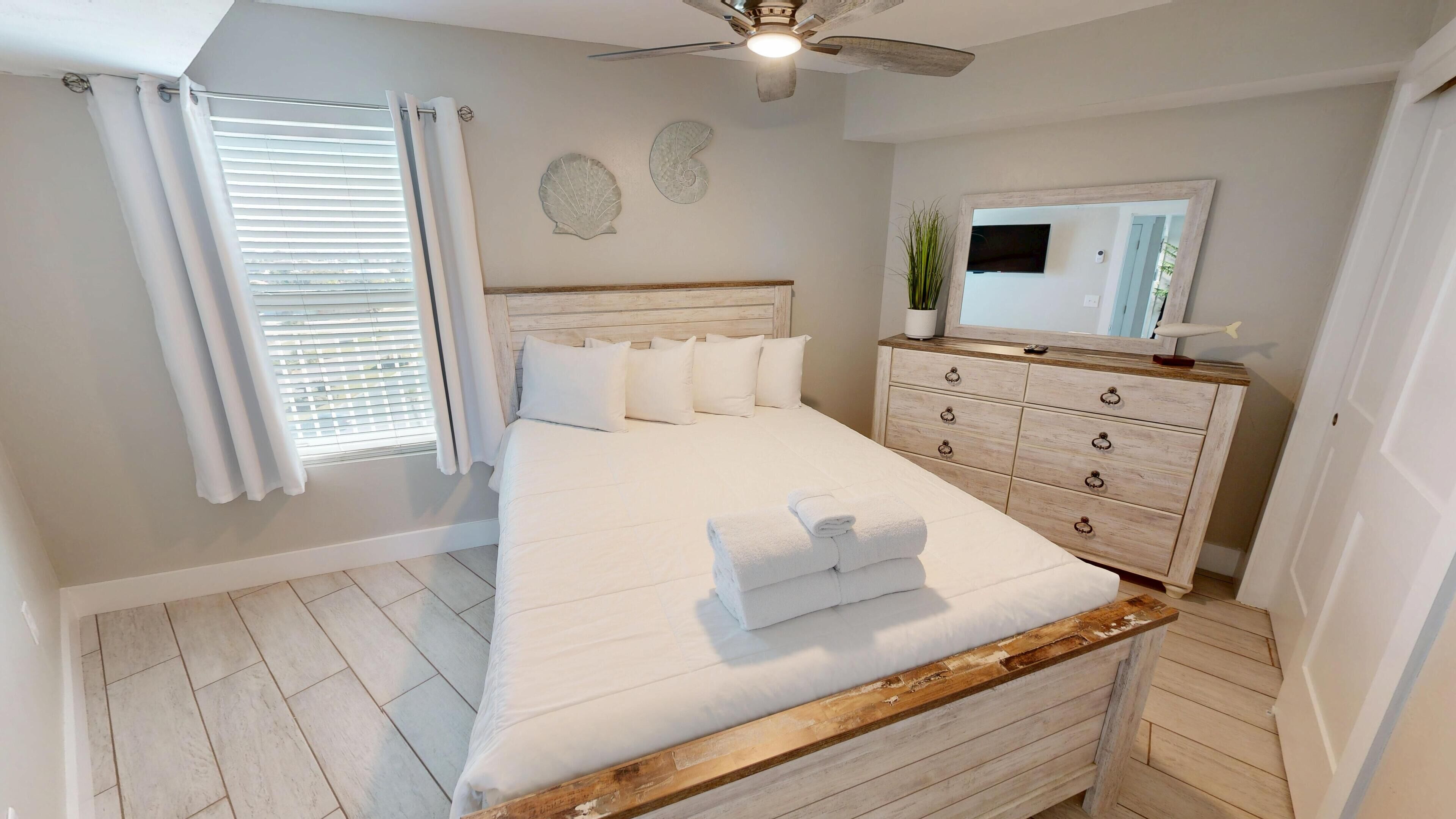 A neatly arranged bedroom with a bed, a dresser, a ceiling fan, and a window with blinds.