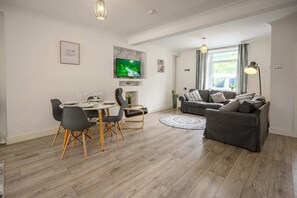 A modern living room with a grey sofa, a dining table with chairs, a flat-screen TV mounted on the wall, and a large window with curtains.