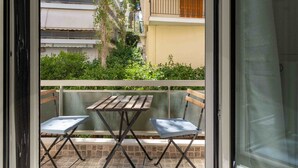 Un balcon avec une table et deux chaises, une vue sur la verdure et les bâtiments, et une porte dotée d’un panneau en verre.