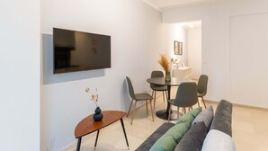 Un salon moderne doté d’un téléviseur à écran plat fixé au mur, d’un coin repas avec une table ronde et des chaises, et d’un canapé agrémenté de plusieurs coussins.