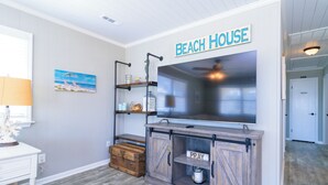 Ett vardagsrum med en stor platt-TV, ett träskåp med en skylt med texten ”BE”, en hylla med dekorativa föremål och en skylt med texten ”STRANDHUS” på väggen.