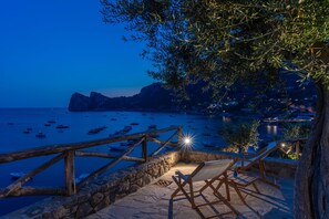 Terraza de piedra con barandillas de madera que da vista a una zona costera con barcos y un pueblo lejano.