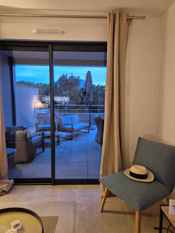 A modern hotel room with a balcony, a grey armchair, a small table, and a hat on the armchair. - Appartement Neuf Élégant à Agosta Plage - Votre Évasion Soleil et Mer (ALBITRECCIA - Agosta)