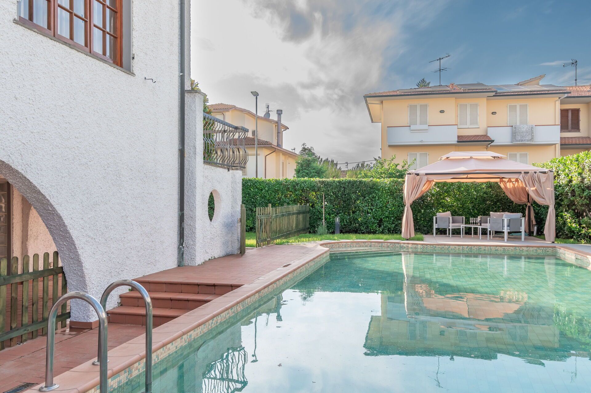 A swimming pool with a pergola and outdoor seating, surrounded by buildings and a hedge.