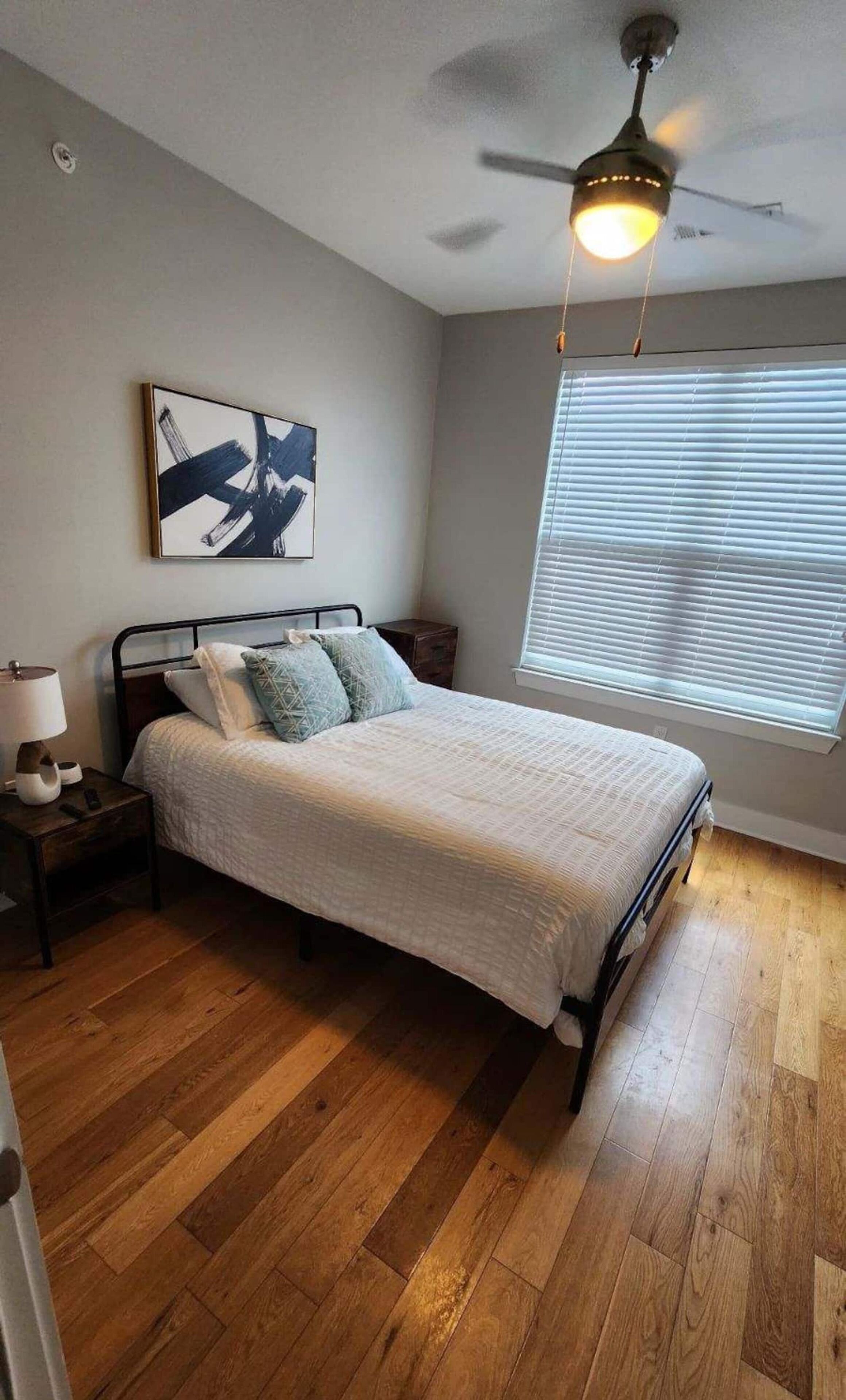 A bedroom with a bed, a nightstand, a ceiling fan, a window with blinds, and a painting on the wall.