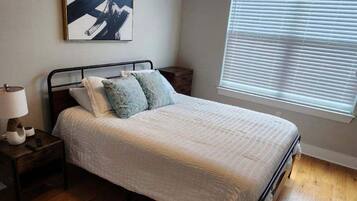 A bedroom with a bed, a nightstand, a ceiling fan, a window with blinds, and a painting on the wall.