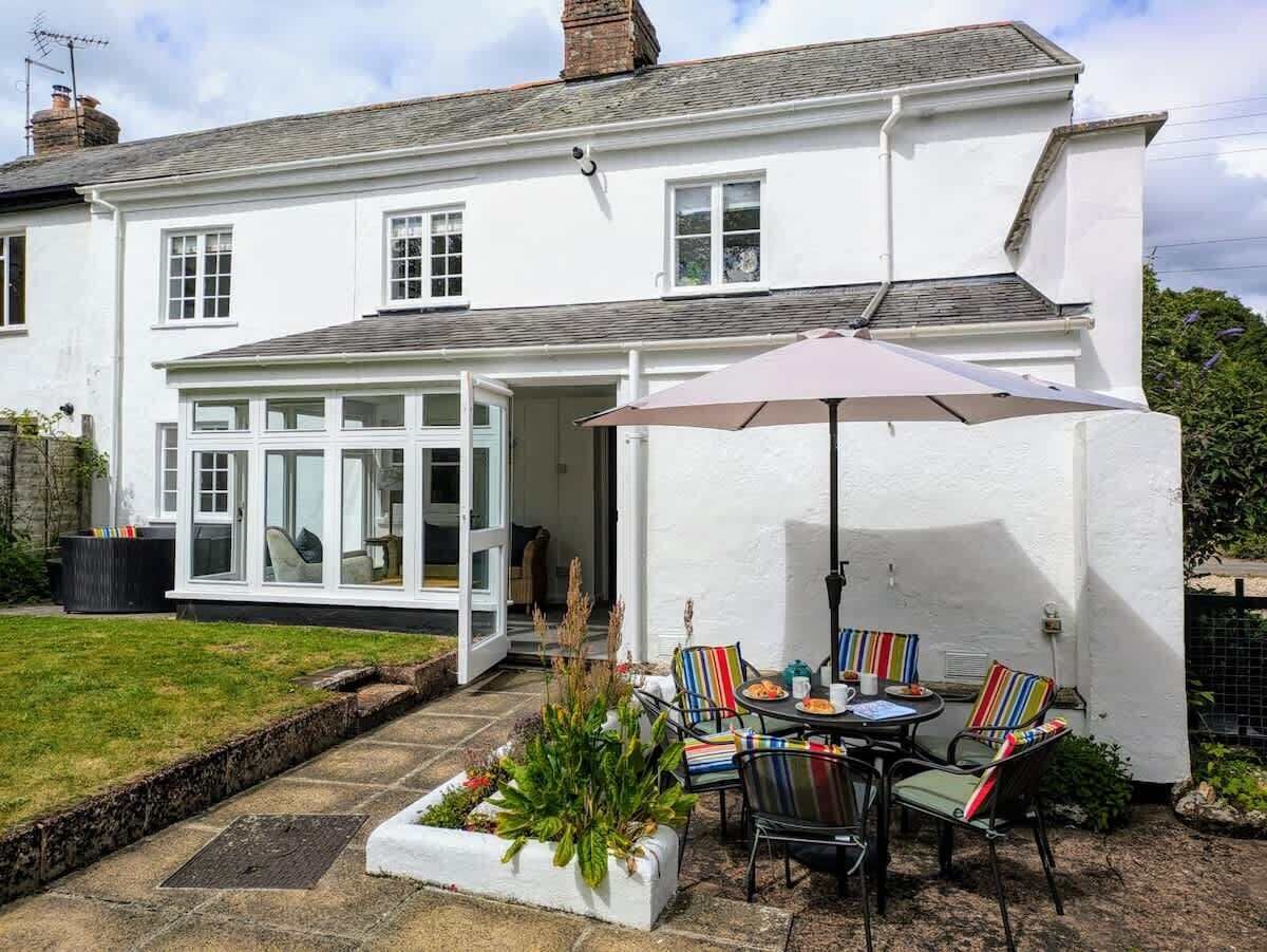 A two-story white house with a patio area featuring a table and chairs, a large umbrella, and a garden.