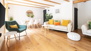Een moderne woonkamer met houten balken, een witte bank met kleurrijke kussens, een groene fauteuil en een eethoek met witte stoelen en een houten tafel.