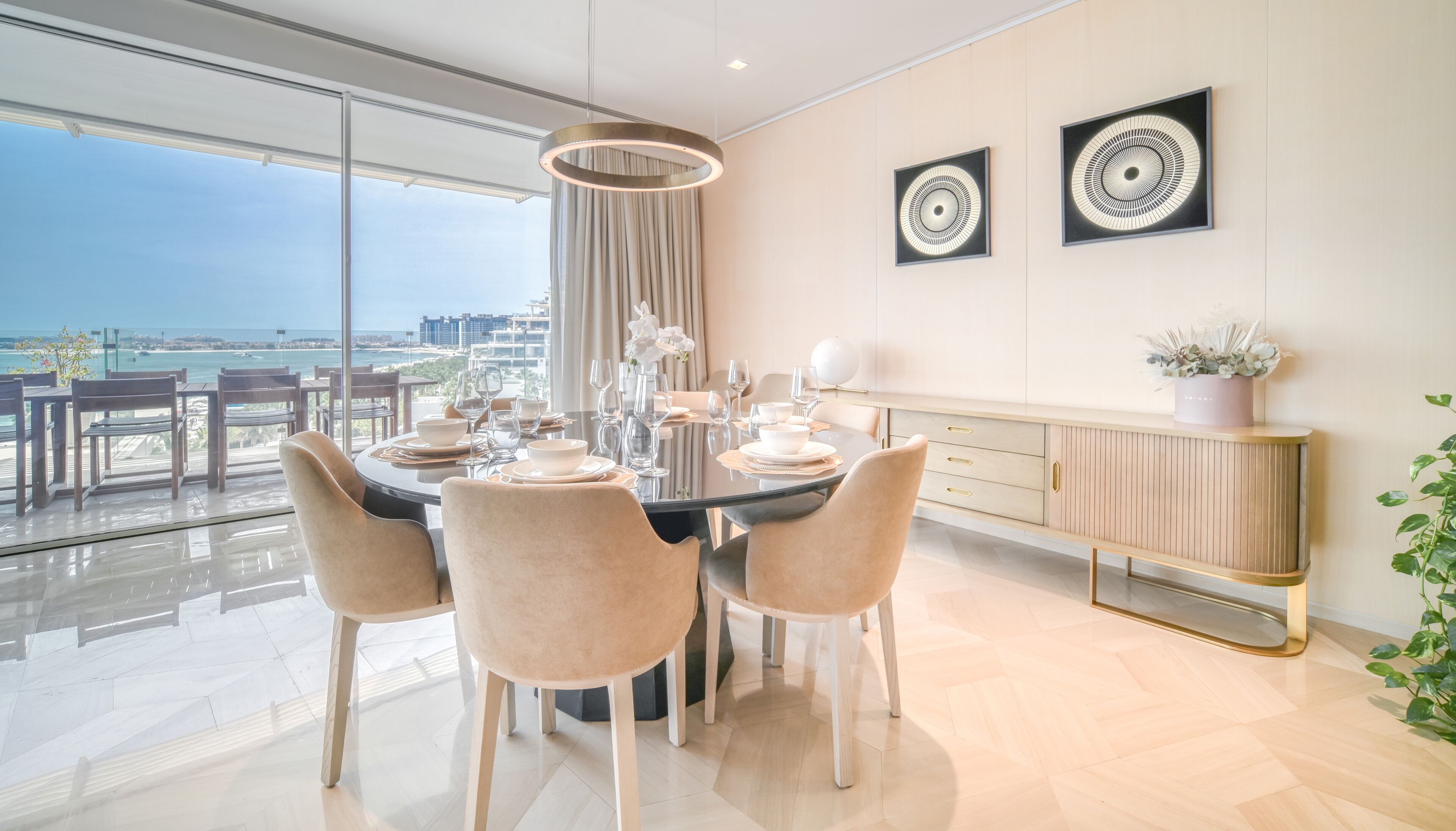 Un espace repas moderne avec une table ronde dressée pour six personnes, de grandes fenêtres offrant une vue sur la mer et un décor minimaliste agrémenté de deux œuvres d'art abstraites accrochées au mur.