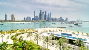 Eine Küstenstadtlandschaft mit einer markanten Skyline, einer Marina mit Yachten und einem Strandabschnitt mit Palmen.