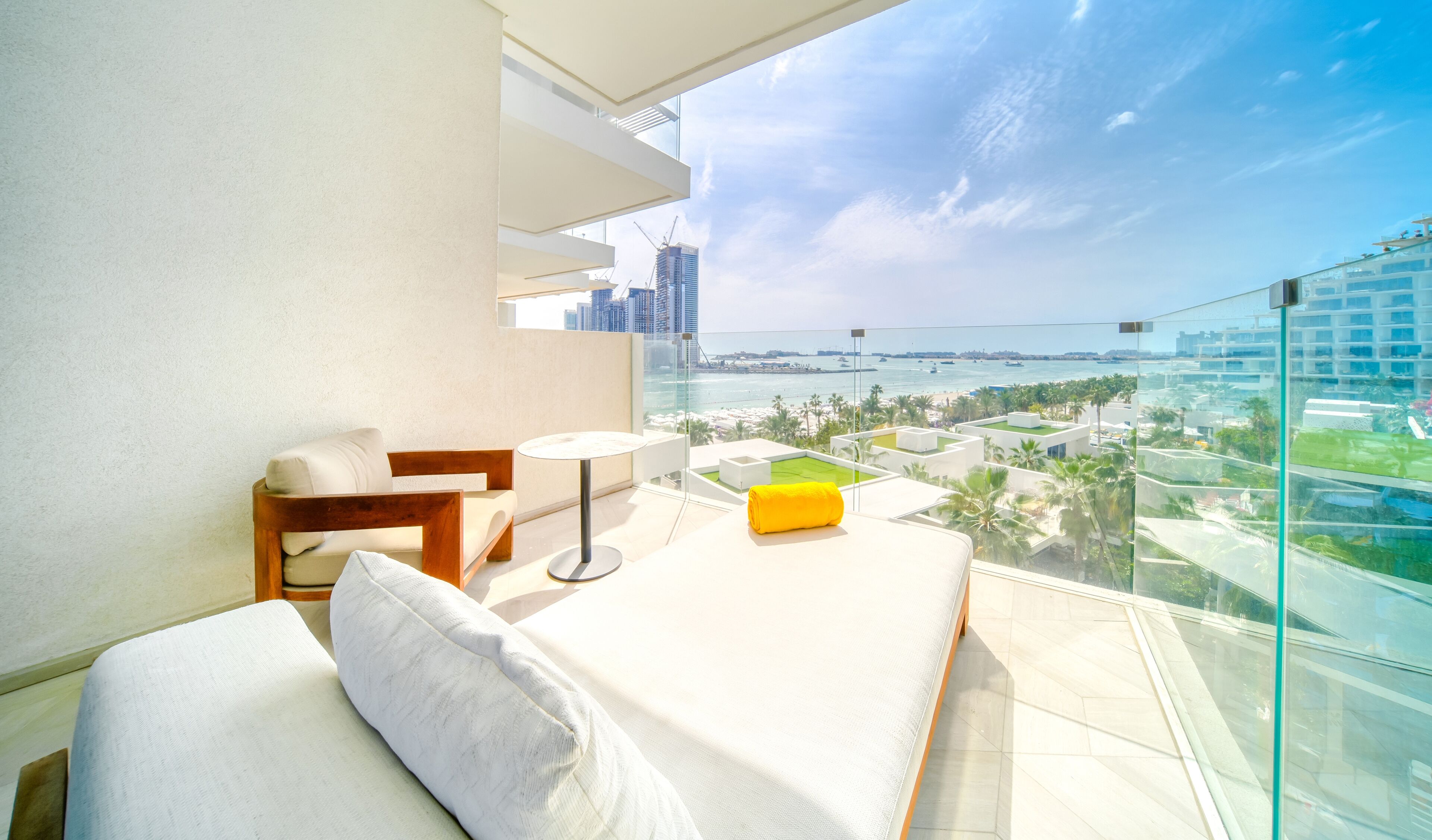 A balcony with a sofa, a small table, and a yellow towel. It overlooks a cityscape and a body of water.