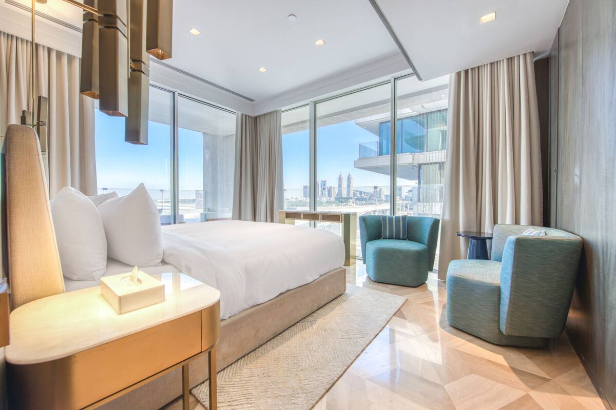 A modern hotel room with a large bed, a seating area with two chairs, and a view of the city skyline through large windows.