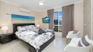 Ein Schlafzimmer mit einem Bett, einem Fernseher, einem Deckenventilator, einem Fenster mit Vorhängen und einem Sessel.