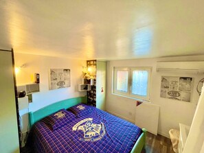 A bedroom with a bed, a wall-mounted shelf with books, a window, and a wall with posters.