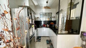 A modern kitchen with white cabinets, a refrigerator, and a microwave. There is a decorative plant in a patterned pot and a wooden chair with a cushion. - Les Pieds Dans L'eau (Valras-Plage)