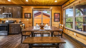 A wooden cabin interior with a dining table set for four, kitchen cabinets, and large windows offering a view of the outdoors.