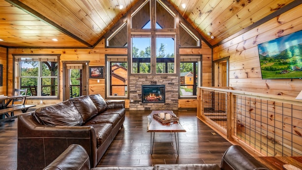 A log cabin interior with a stone fireplace, leather sofas, and large windows offering views of the surrounding forest.