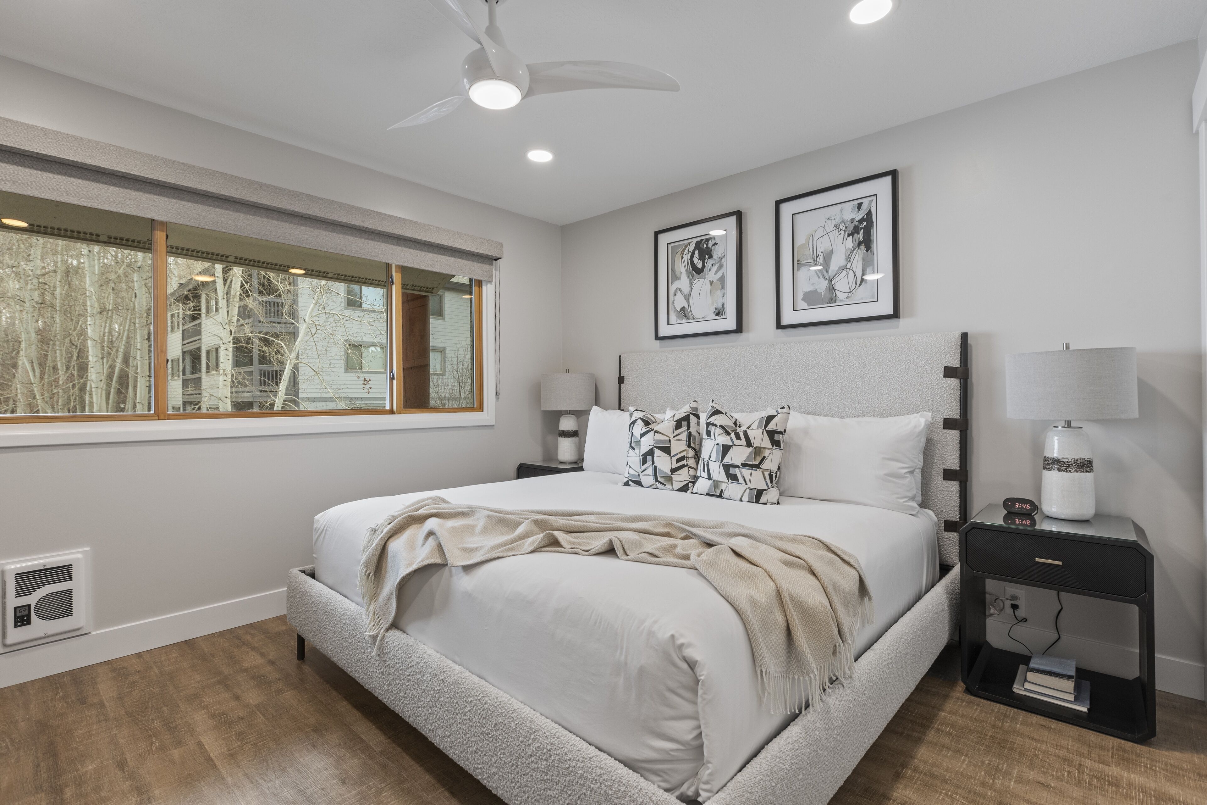 A bedroom with a large bed, a window, a ceiling fan, and two framed pictures on the wall.