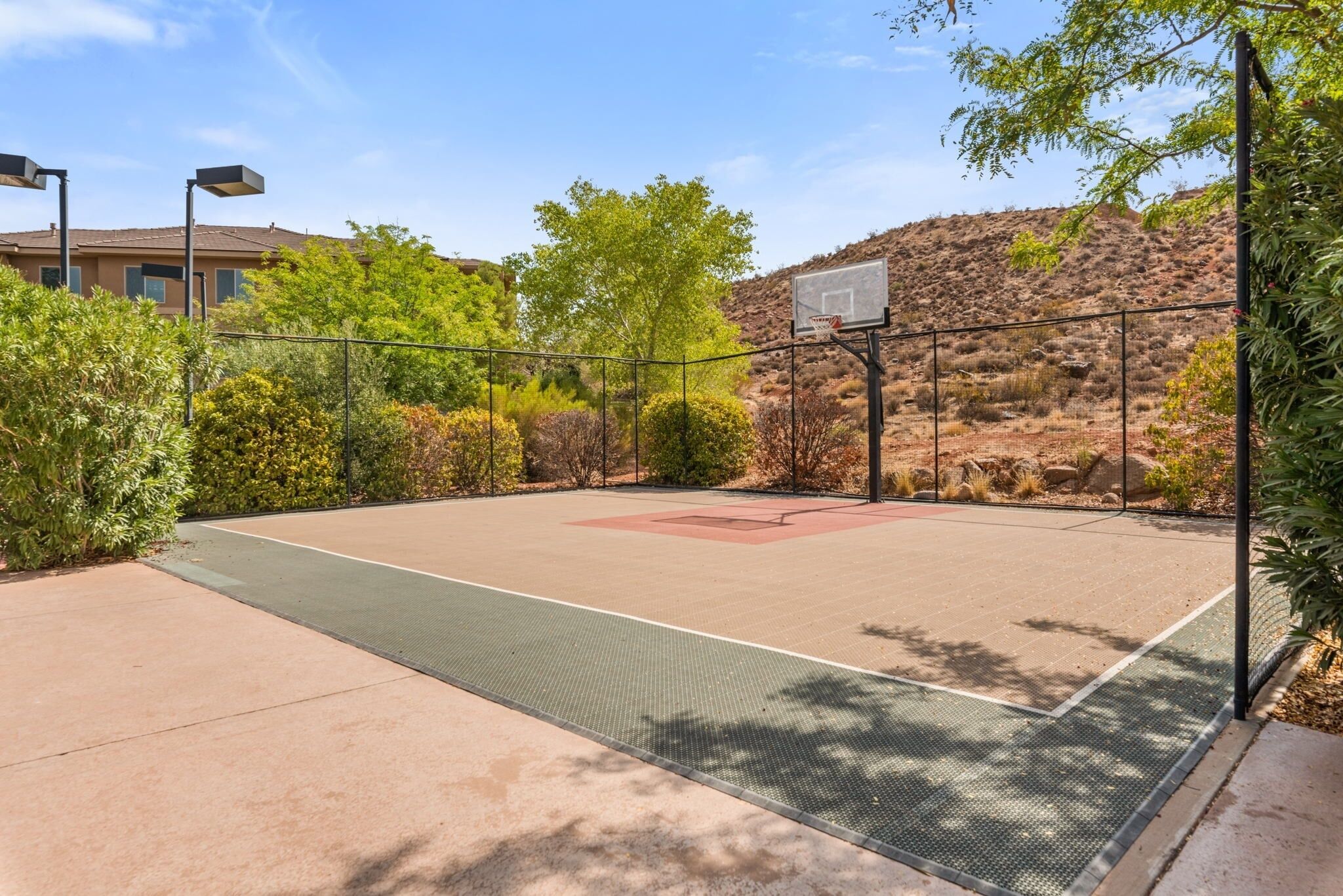 An outdoor basketball court with a metal fence, trees, and a building in the background.