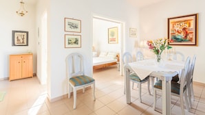 A dining area with a table set for four, surrounded by chairs with cushioned seats. There is a wooden cabinet to the left and a bed with a white headboard in the background. - Casa Nebari Seaside Home-fully Equipped Ideal Spot (Messini)