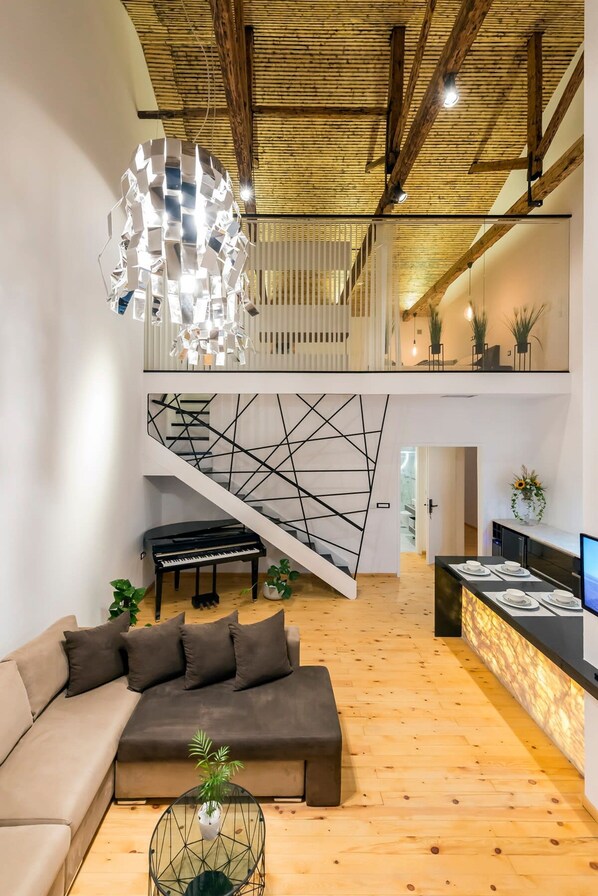 A modern interior with a wooden ceiling, a staircase with geometric wall art, a piano, a sofa, a dining area, and a hanging light fixture.