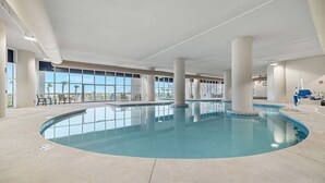 Une piscine intérieure à l’eau d’un bleu cristallin, entourée de grandes fenêtres qui offrent une vue sur des palmiers et un ciel dégagé.