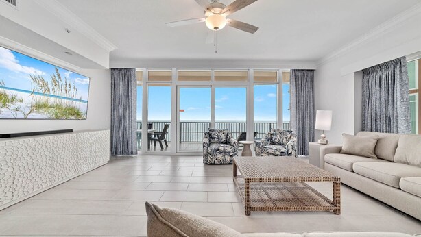 Un salon spacieux doté d’une grande fenêtre, d’un ventilateur de plafond et d’un balcon offrant une vue sur la plage.
