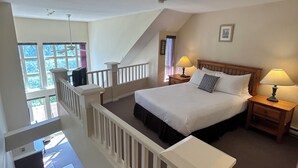 A bedroom with a large bed, a wooden headboard, two bedside tables with lamps, a window with curtains, and a staircase leading to an upper floor. - Classic Loft - 309 (Kelowna)