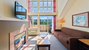 A living room with a fireplace, a television, a sofa, a table, and a balcony with a view. - Classic Loft - 309 (Kelowna)