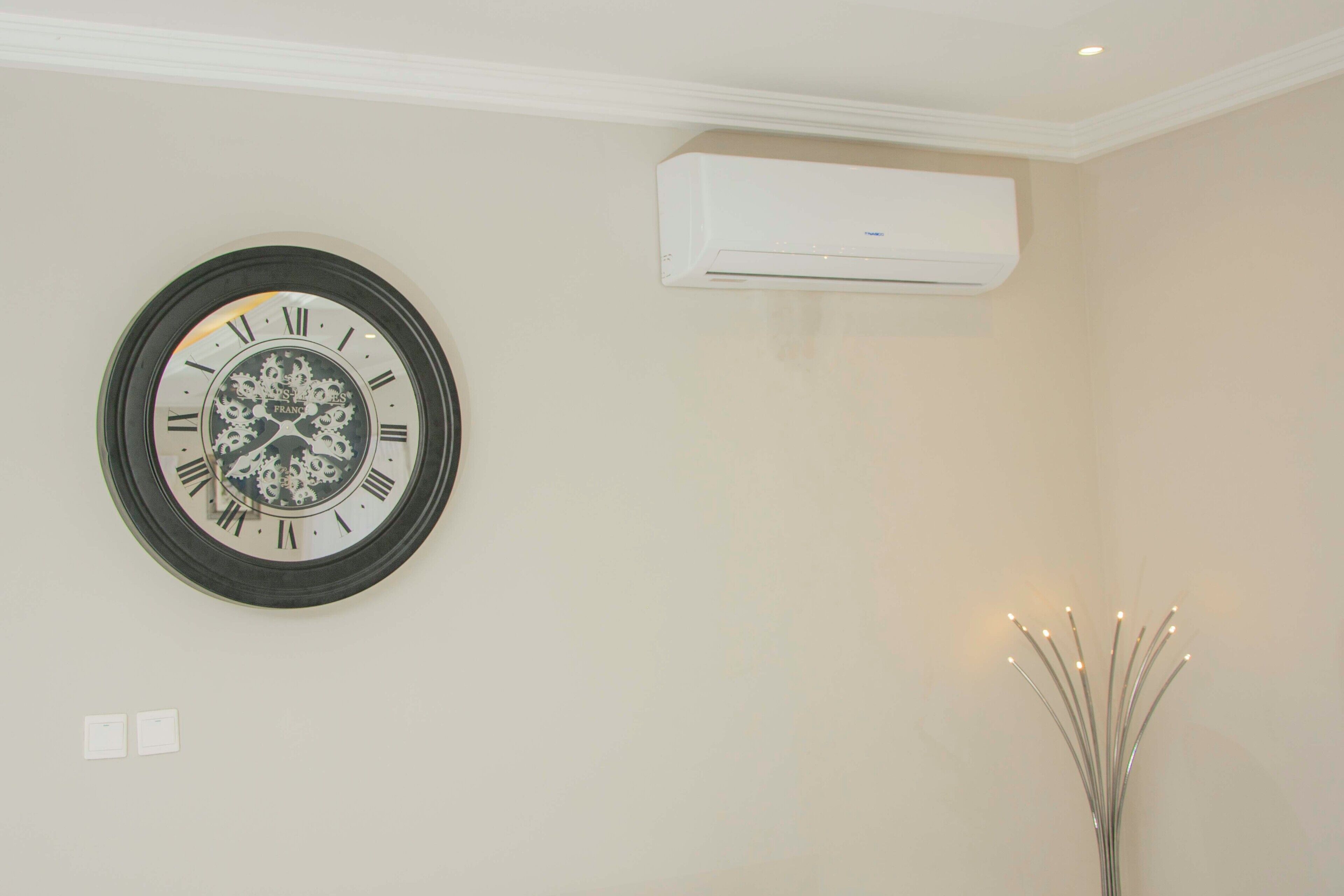 Een hangend klok met Romeinse cijfers en decoratieve details, een airconditioning en een eenvoudige lamp.