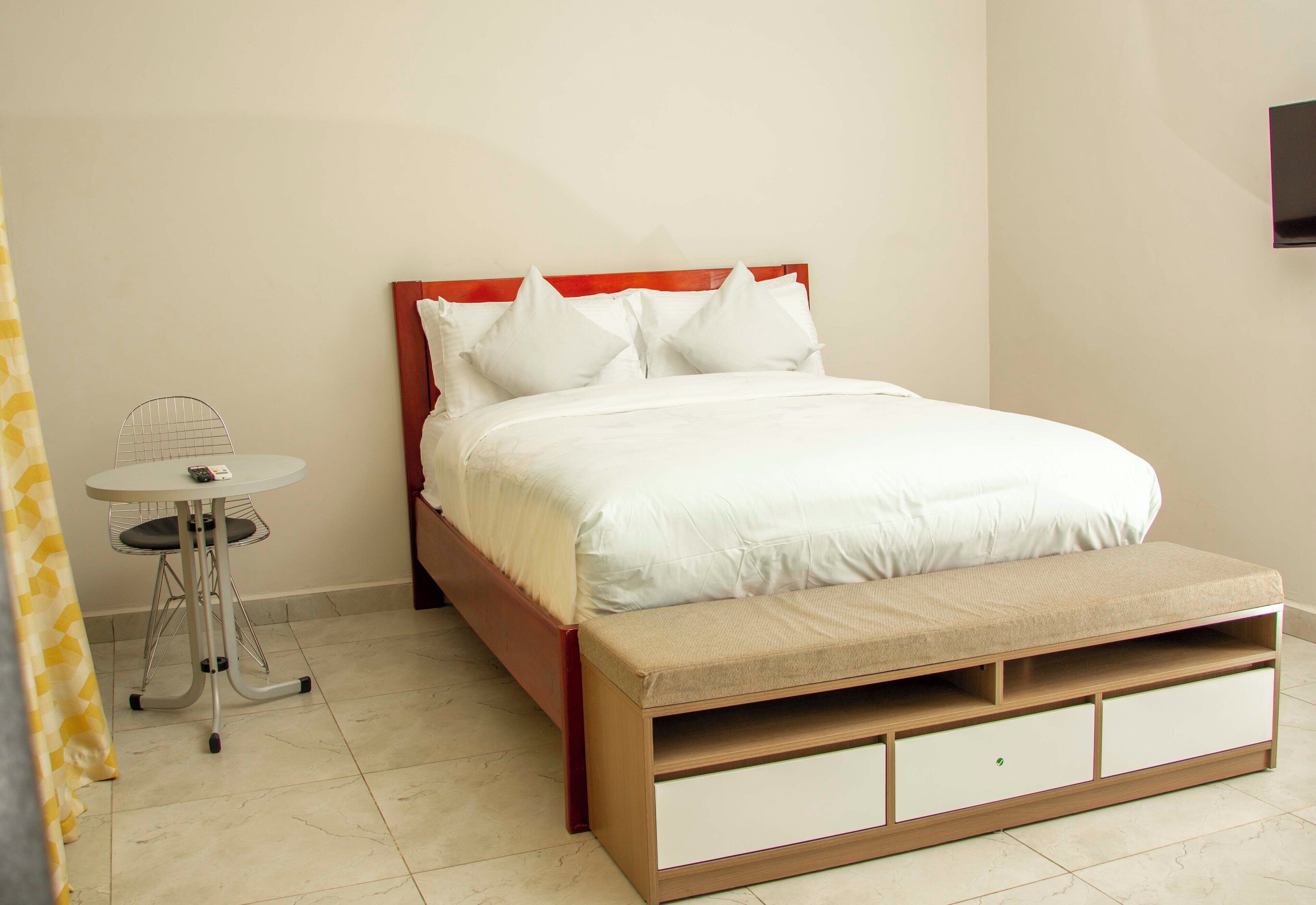 A bed with a wooden frame and white bedding, a small table with a chair, and a bench with storage drawers.