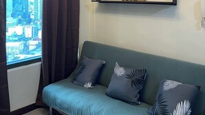 A modern living room with a green sofa, decorative pillows, a city view through a large window, and a ceiling light fixture. - Lovely 1-bedroom condo in fabulous Makati with WiFi, AC, fitness room (Makati)