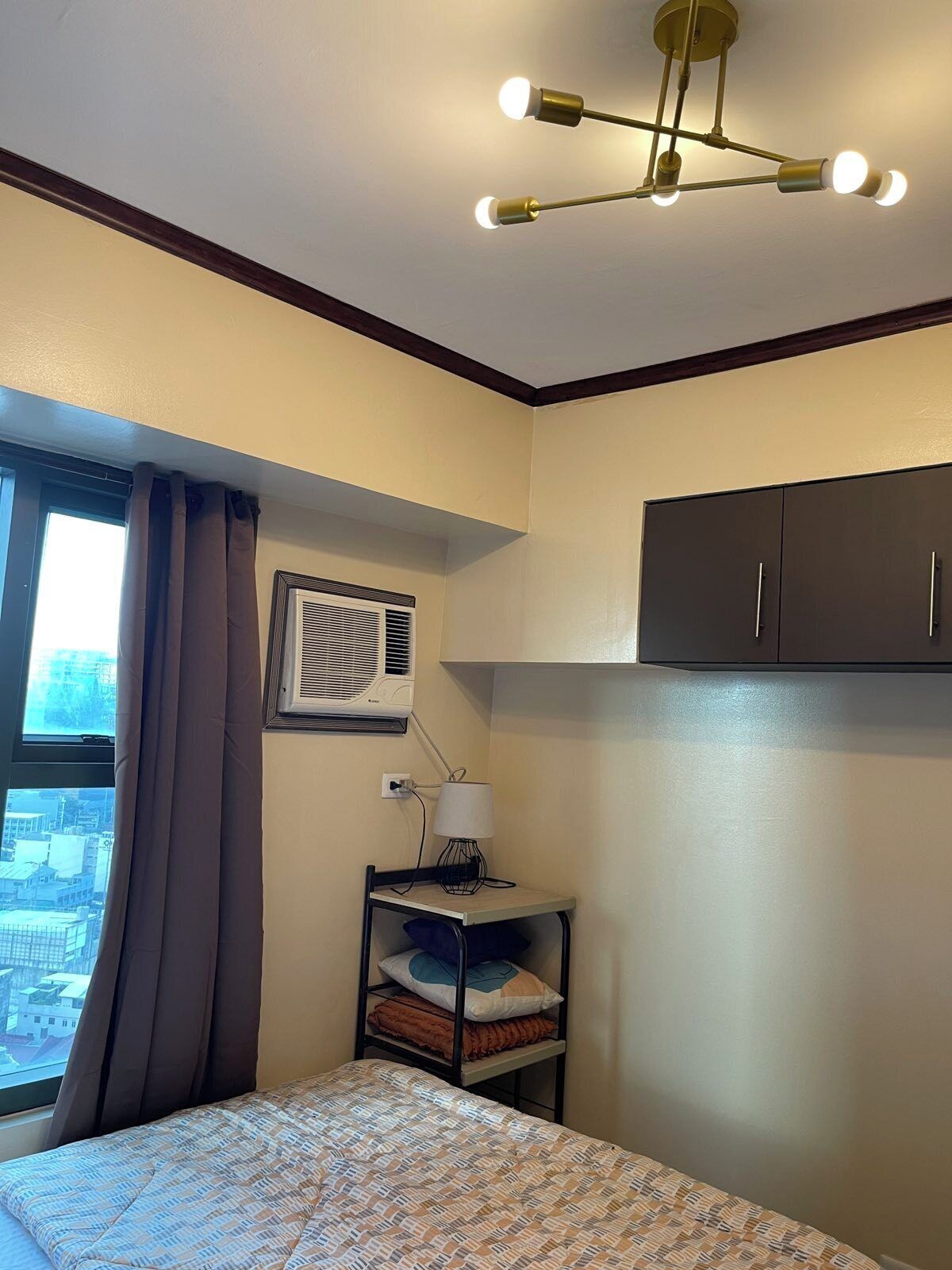 Ein Hotelzimmer mit einem Bett, einem Schreibtisch mit Lampe, einem Fenster mit Vorhängen, einer Klimaanlage und einer Deckenleuchte.