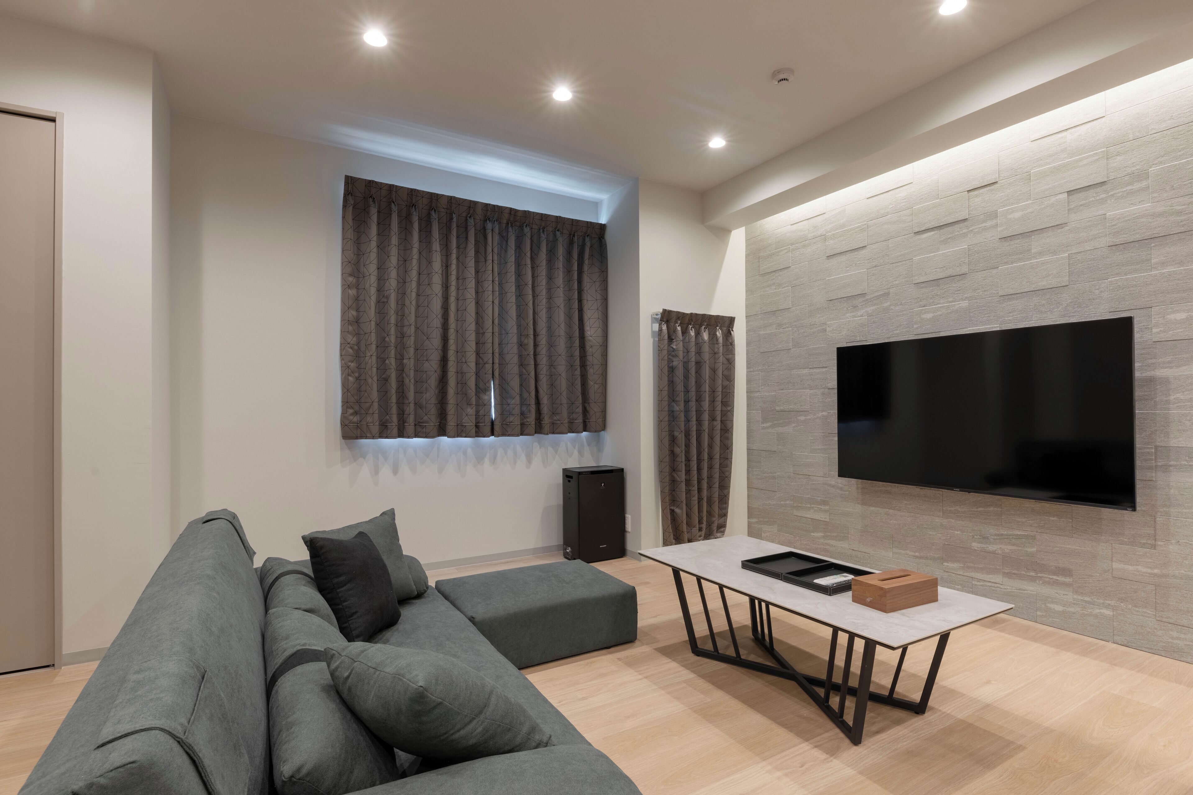 A modern living room with a grey sectional sofa, a flat-screen TV mounted on a grey stone wall, a small table with a box, and a speaker.