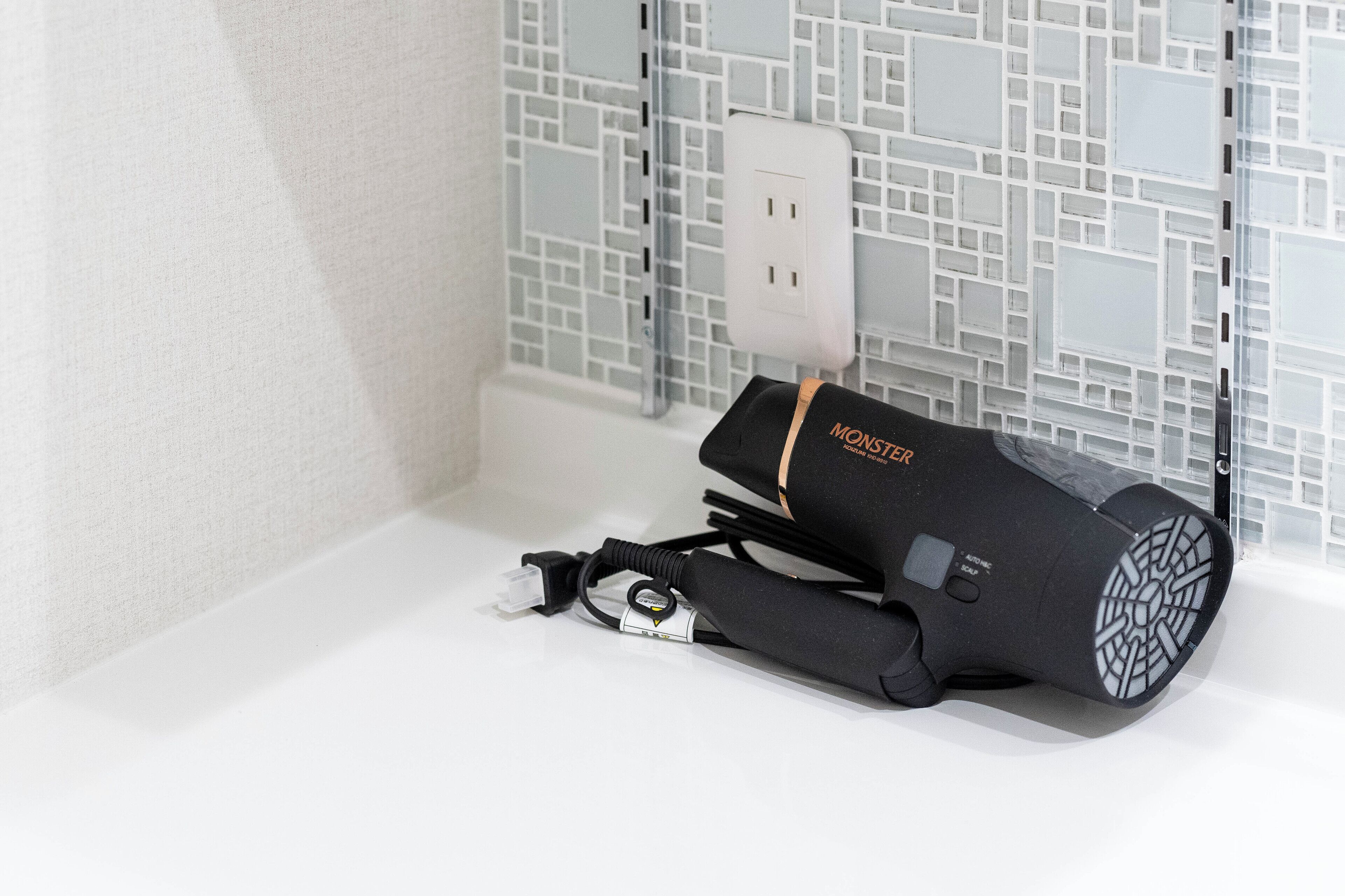 A black portable air purifier with a cord and a plug, placed on a white surface against a tiled wall.