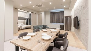 A modern dining area with a long wooden table set for four, surrounded by black chairs. A grey sofa and a bed with a grey headboard are visible in the background. - Crice Hotel Shinsaibashi (Osaka)