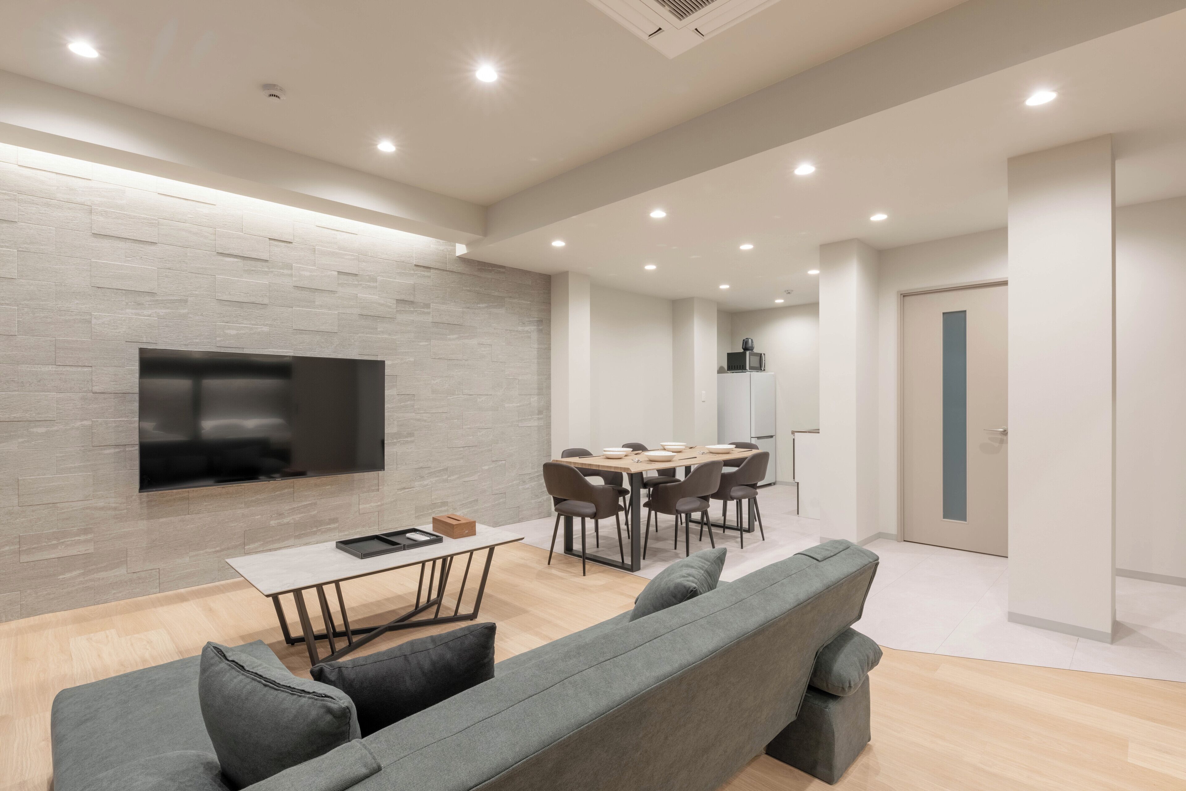 A modern living room with a grey sofa, a dining table with chairs, a flat-screen TV mounted on a textured wall, and recessed ceiling lighting.