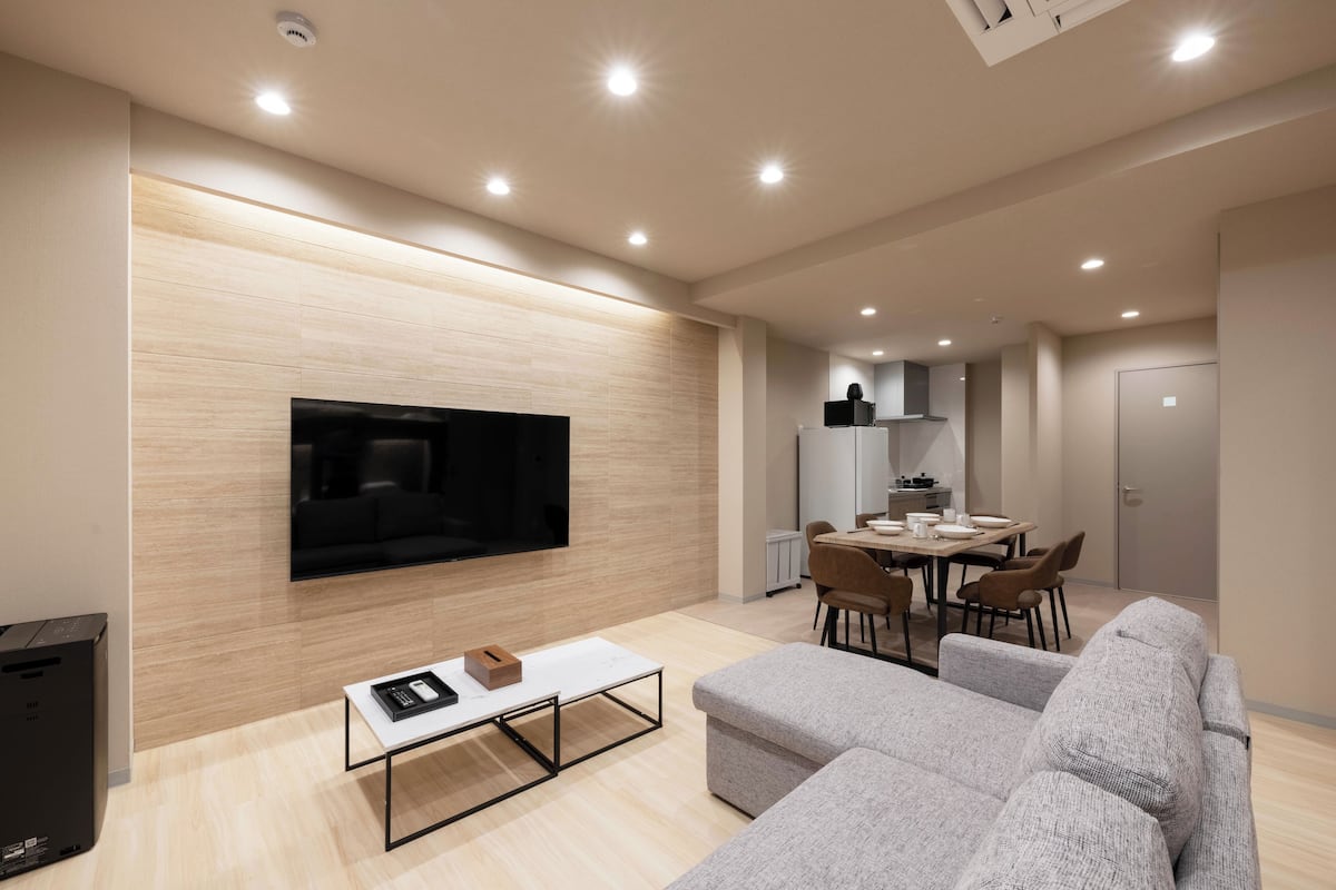A modern living room with a flat-screen TV, grey sofas, a coffee table, and a dining area with chairs and a table.